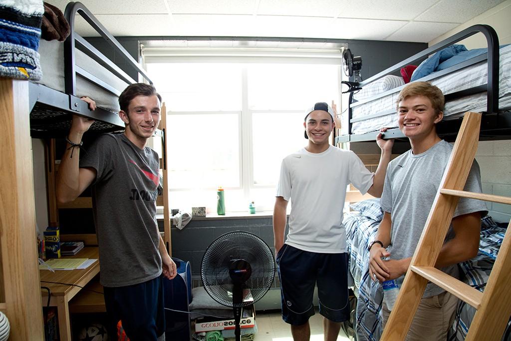 Three U N E students stand near their bunk beds in a dorm room